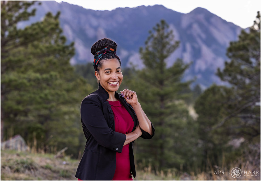 Stunning woman poses in front of the beautiful Boulder mountains in a forest setting while wearing a bun tied on top of her head with a multi colored scarf and a maroon dress and black jacket over it