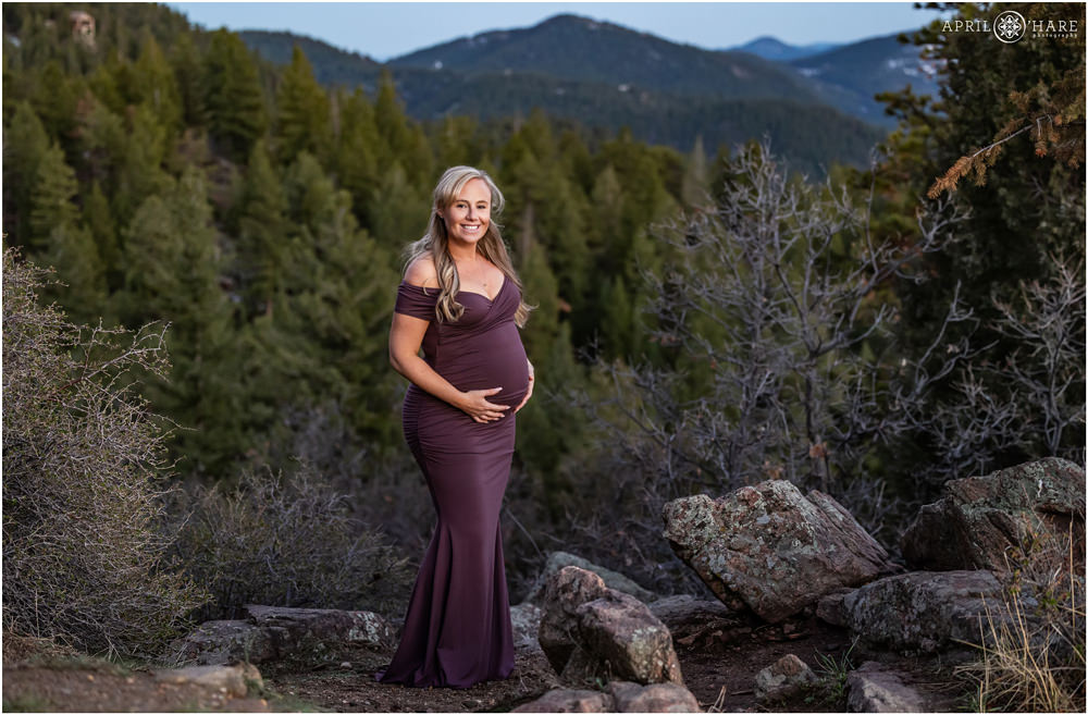 A gorgeous blone woman wearing a long form fitting lilac dress poses in front of a forested mountain view in Colorado