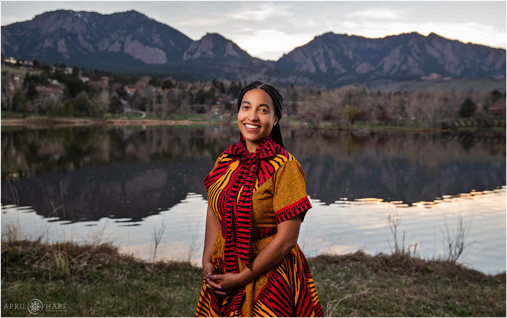 Woman with a beautiful smile and long braided hair holds her hands at her waist while wearing a cool red and yellow orange patterened dress in front of Viele Lake in Boulder Colorado