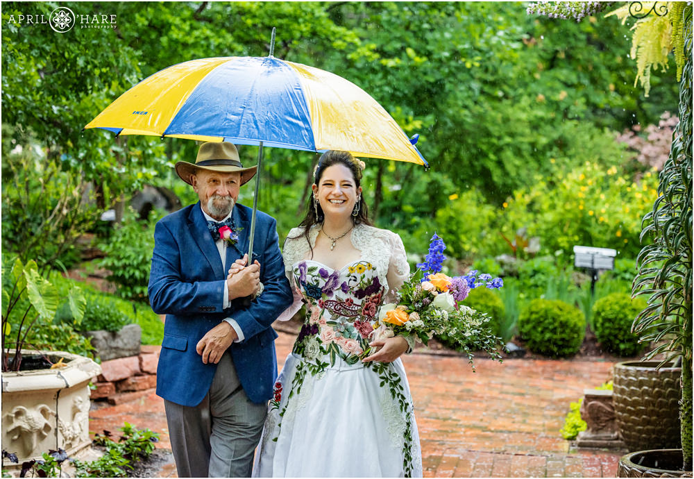 Bride walks with her day with pouring rain coming down under an umbrella at Woodland Mosaic Garden