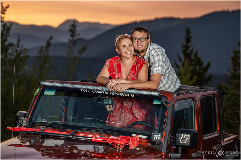 Couple pose standing through the open roof of their red Jeep in front of a pretty Colorado mountain sunset backdrop