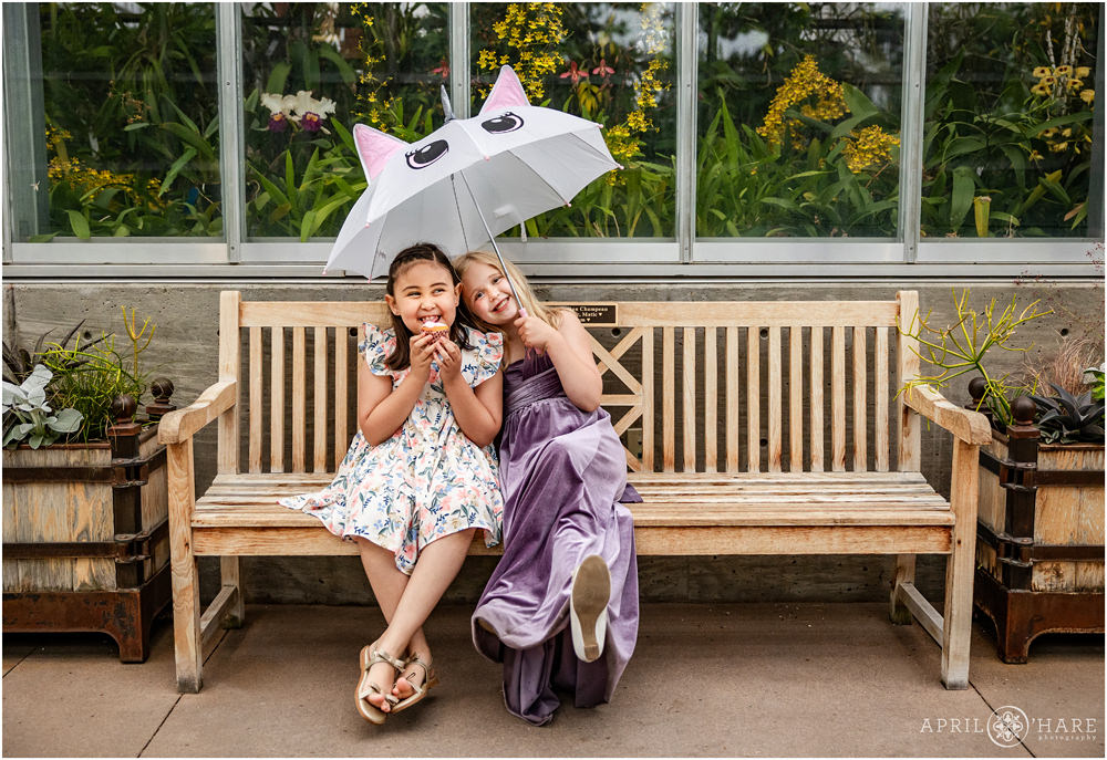 Sweet photo of two girls at a wedding sitting on a bench with an umbrella eating a cupcake at the Orangery wedding reception at Denver Botanic Gardens