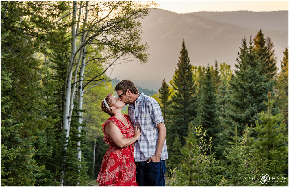 A couple kiss in front of a pretty forested trail with a mountain backdrop at sunset