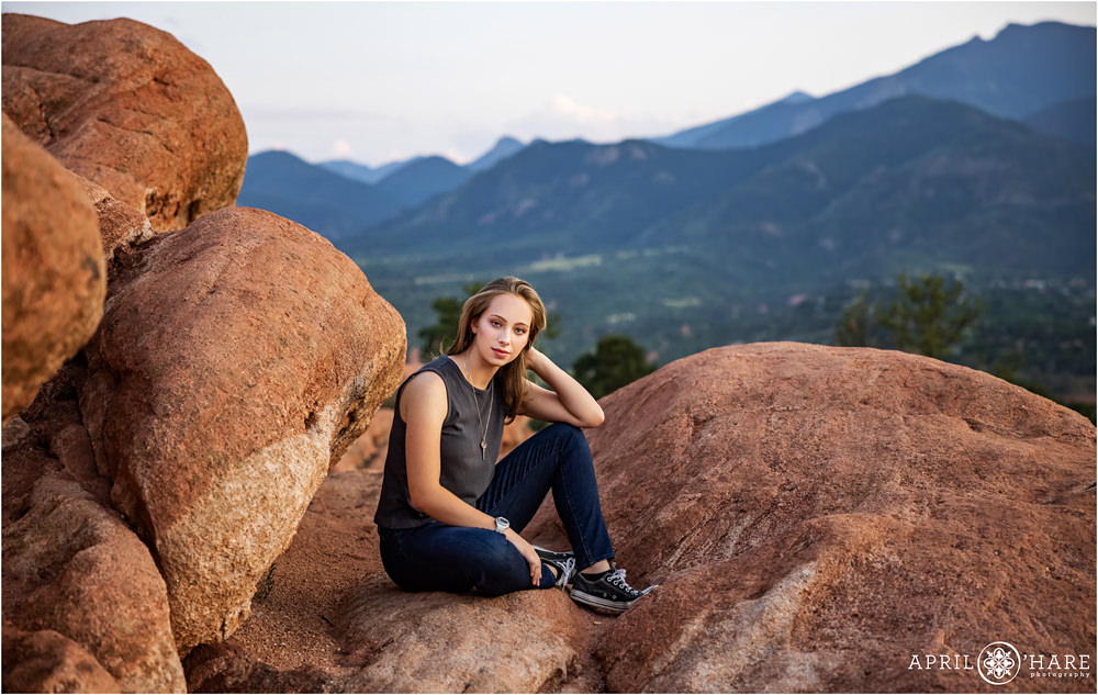 High school senior girl wearing jeans and a tank top sits on a red rock at Garden of the Gods with blue mountains in the backdrop in Colorado Springs