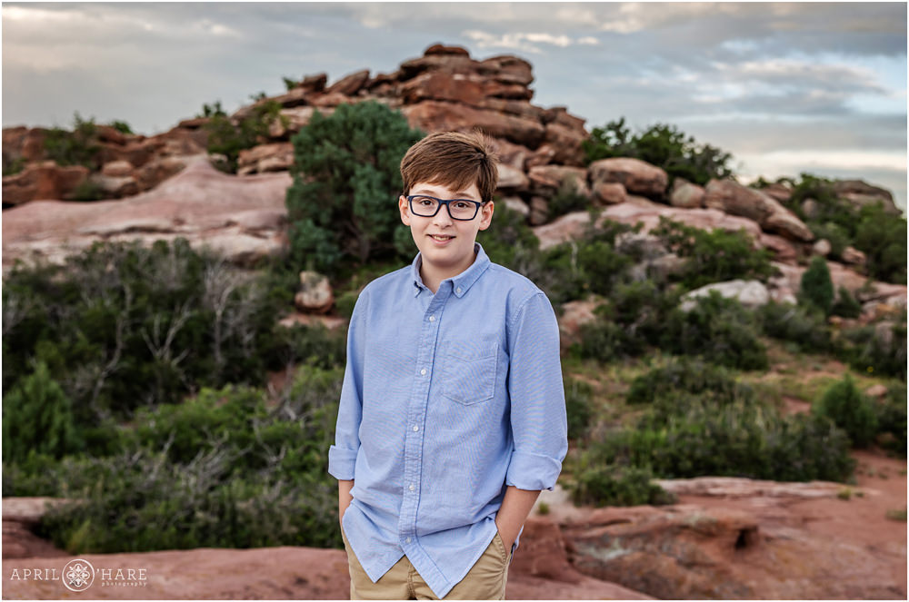 Young boy wearing glasses and light blue shirt smiles for an individual photo in front of red rocks backdrop in Ken Caryl