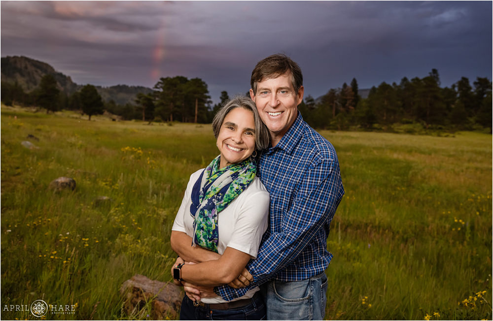 A pretty rainbow and dramatic stormy sky and a wildflower field make up the backdrop for this pretty evening photo of a couple at Rocky Mountain National Park
