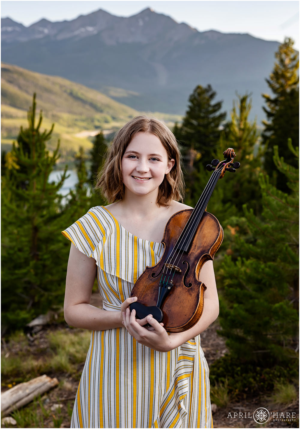 Teenage girl wearing a striped summer dress poses for a portrait with her violin at Sapphire Point in Colorado