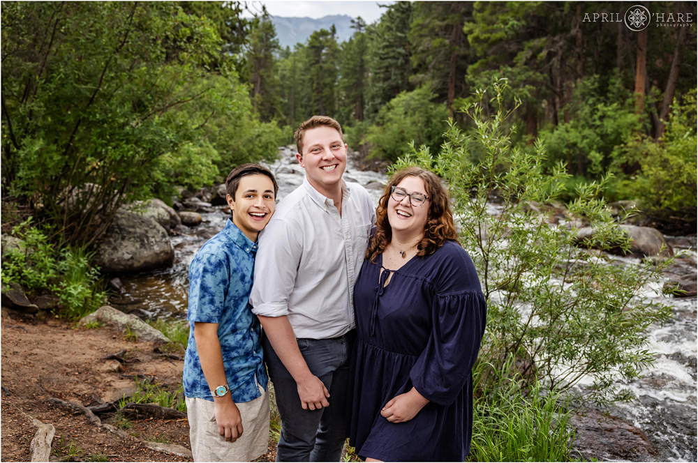 3 siblings laugh together with the Big Thompson River as their backdrop at their family's Estes Park photography session in Colorado