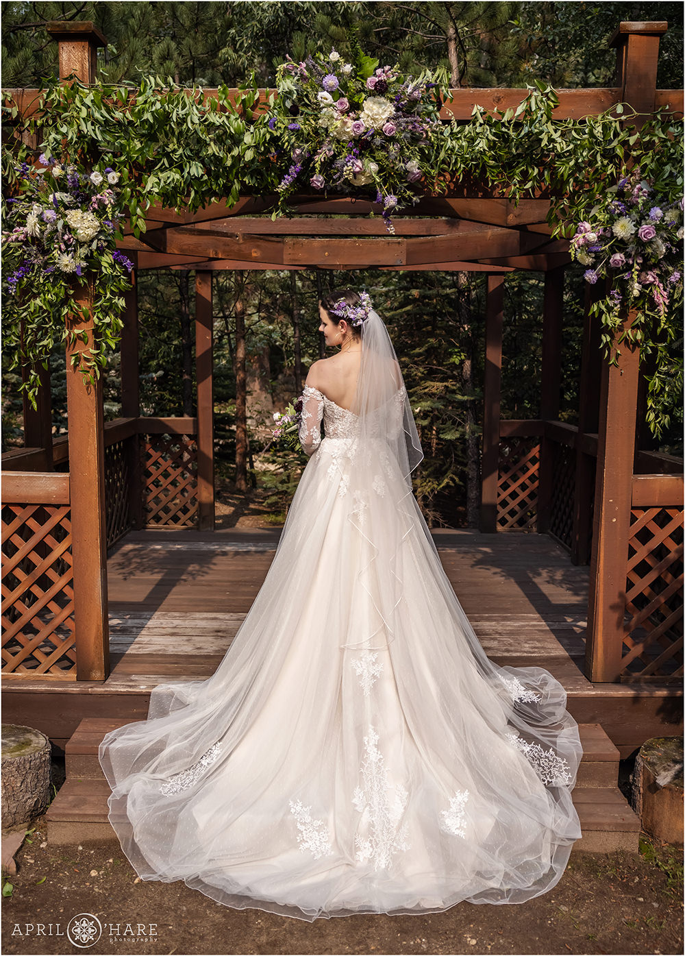 Bride stands underneath wood gazebo decorated with purple flowers from Best Day Floral with the back of her dress toward the camera
