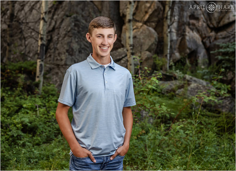 High school senior boy wearing a light blue shirt and jeans poses in the woods in Colorado