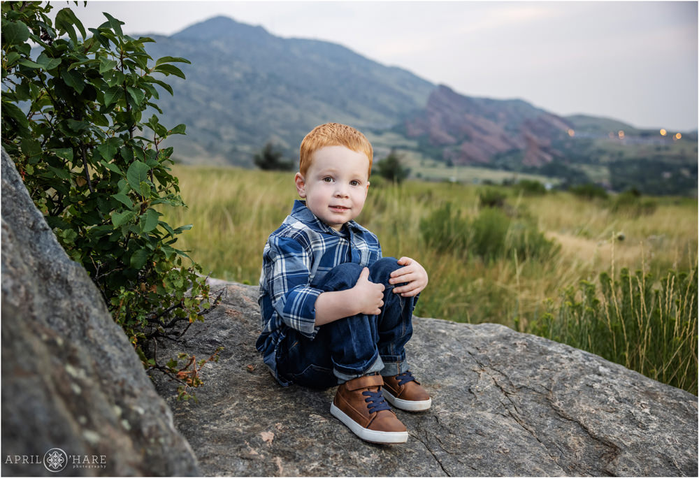 Cute little redhead boy wearing blue plaid and brown shoes with blue laces sits on a rock at East Mount Falcon Trailhead in Morrison Colorado