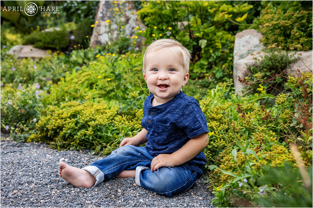 Adorable one year old baby boy wearing blue sits in front of a pretty green garden at Denver Botanic Gardens