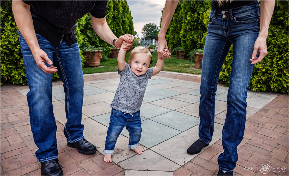 Sweet one year old boy smiles as he holds his parents hands in front of the Romantic Gardens area of Denver Botanic Gardens