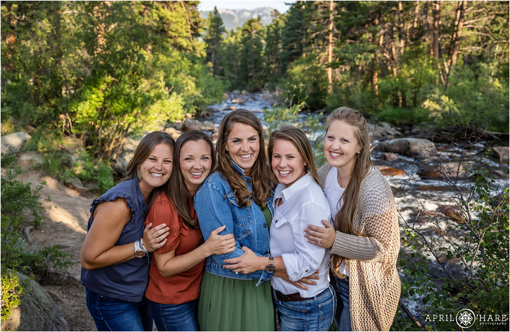 5 adult sisters laugh together in front of a pretty river scene inside Rocky Mountain National Park during summer