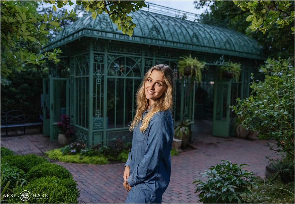 Pretty blonde senior girl wearing a long sleeved blue chambray shirt dress in front of historic green solarium at Denver Botanic Gardens