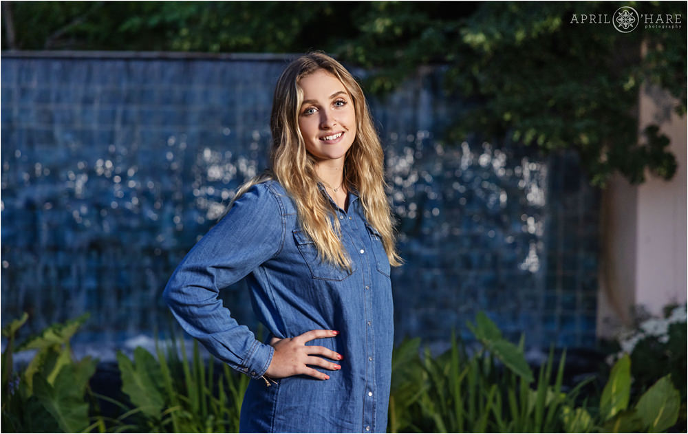 High School Senior girl with long blonde hair wearing a blue chambray shirtdress in front of a pretty blue tile waterfall fountain at Denver Botanic Gardens