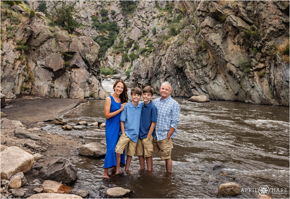 Family of four wearing blue with two young teen sons stand in the Big Thompson River together with the dramatic rocky backdrop at Viestenz-Smith Mountain Park during summer