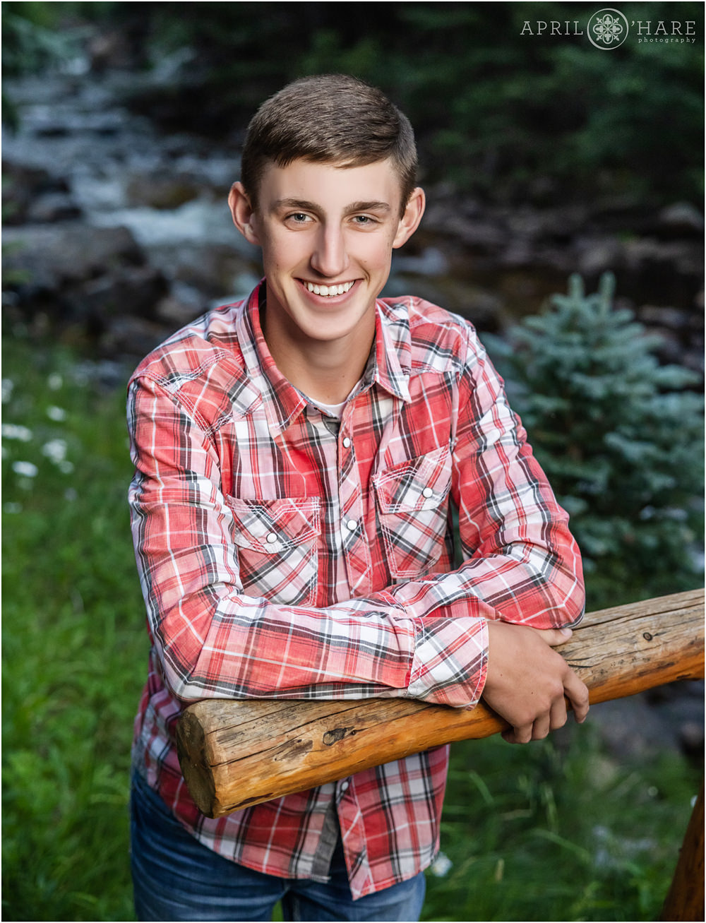 Senior boy wearing a red plaid button down shirt poses in front of a pretty mountain stream in Colorado
