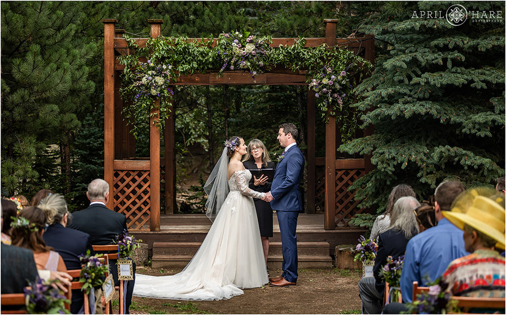 Bride and groom hold hands facing each other under purple floral display attached to a red wood gazebo at Estes Park Condos