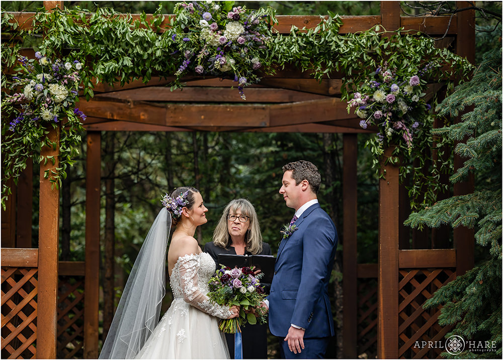 Bride and groom stand under red wood gazebo decorated with purple flowers and greenery on their wedding day outdoors in Colorado