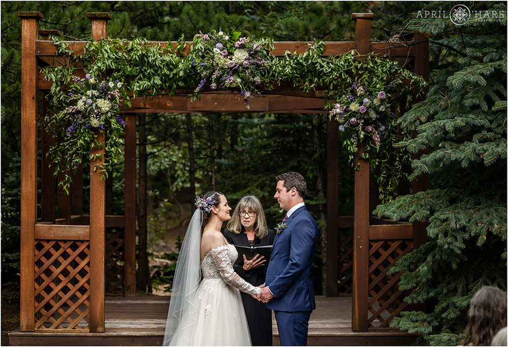 Bride and groom hold hands and look at each other at their outdoor Estes Park Wedding with a wood gazebo backdrop decorated with purple florals from Best Day Floral