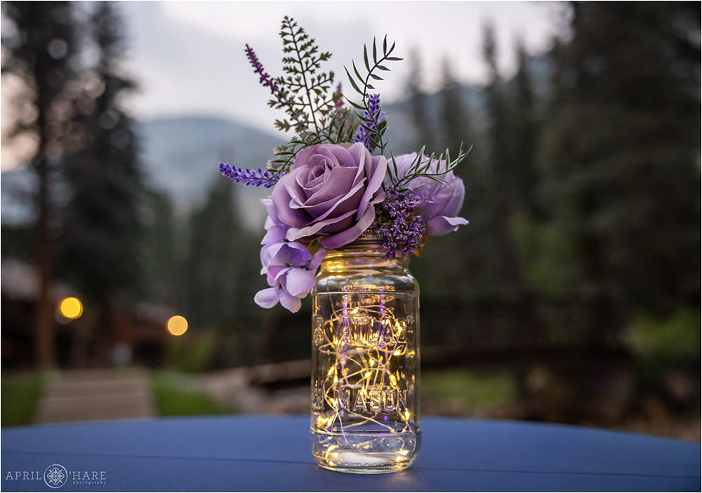 Silk flowers in a mason jar with twinkle lights inside with mountain backdrop on a cocktail table at an outdoor wedding at Estes Park Condos