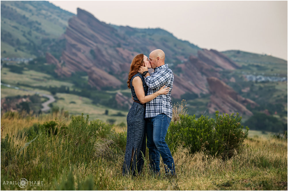 Couple kiss in front of a pretty Red Rocks backdrop at East Mount Falcon Trailhead on a hazy summer day in Colorado
