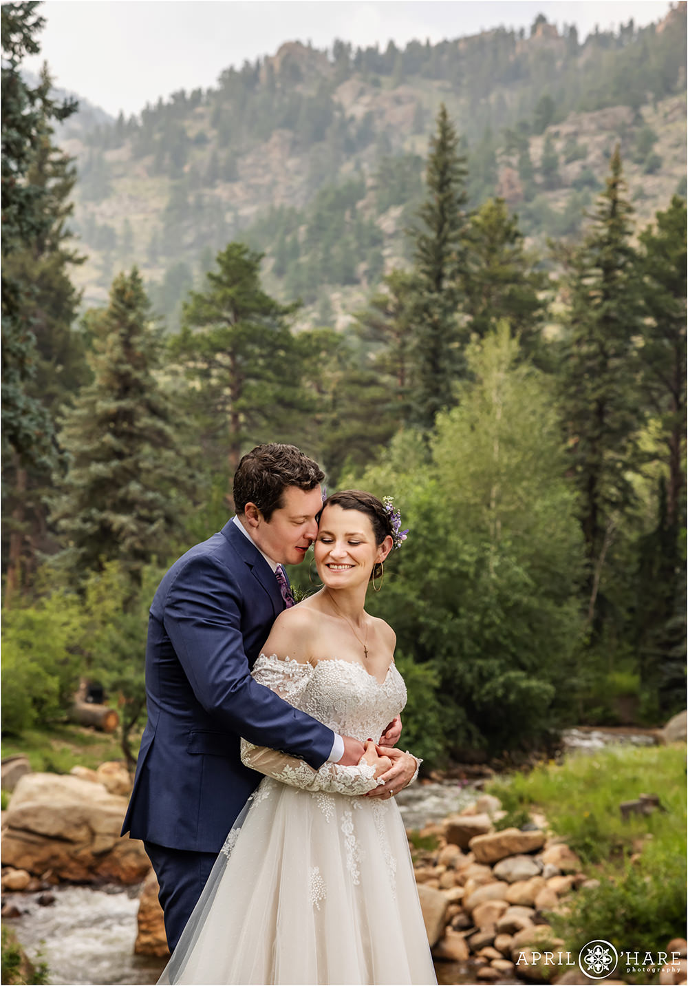 A vertical photo of a bride and groom snuggling with Fall River and mountain backdrop behind them