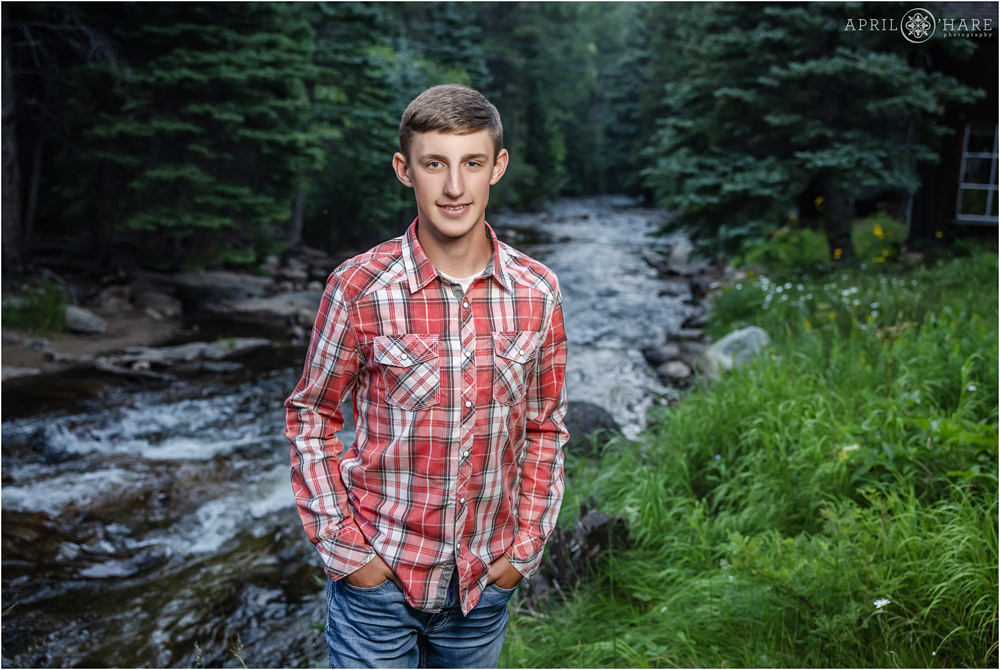 Senior boy wearing a long sleeve plaid button down shirt and blue jeans poses in front of a beautiful mountain stream with wildflowers in the backdrop in Boulder Colorado