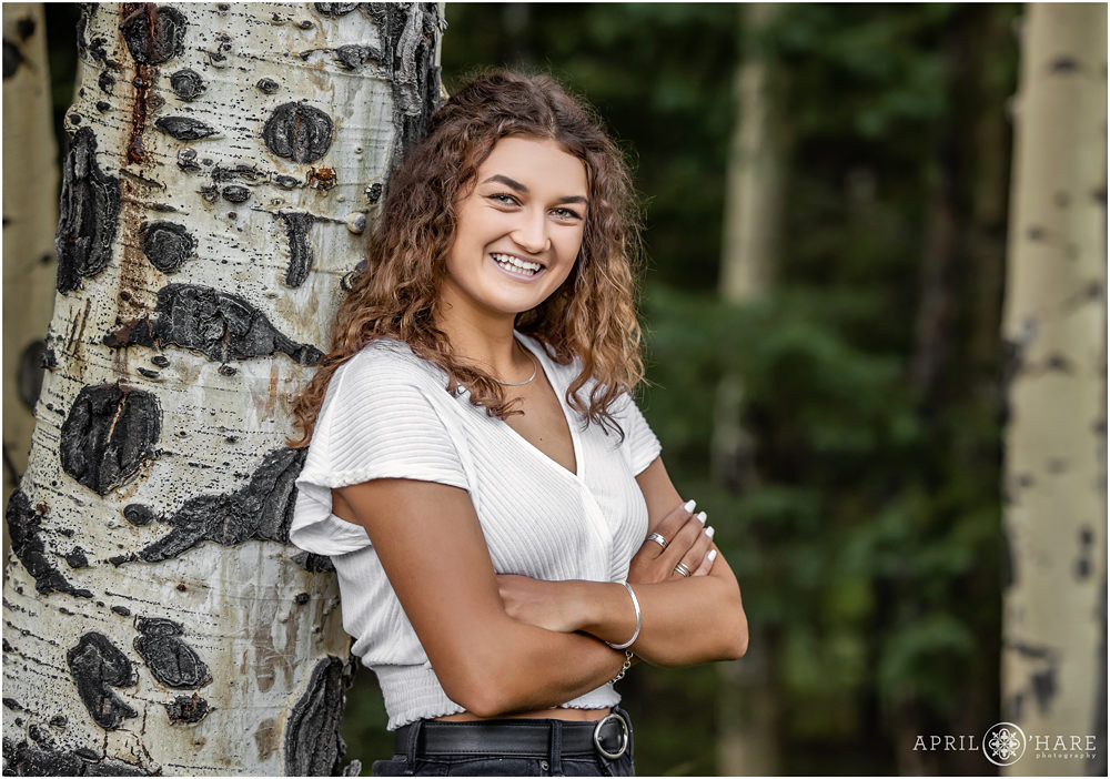 High school senior girl leans against a black and white aspen tree trunk in the woods of Evergreen Colorado for her senior photo session
