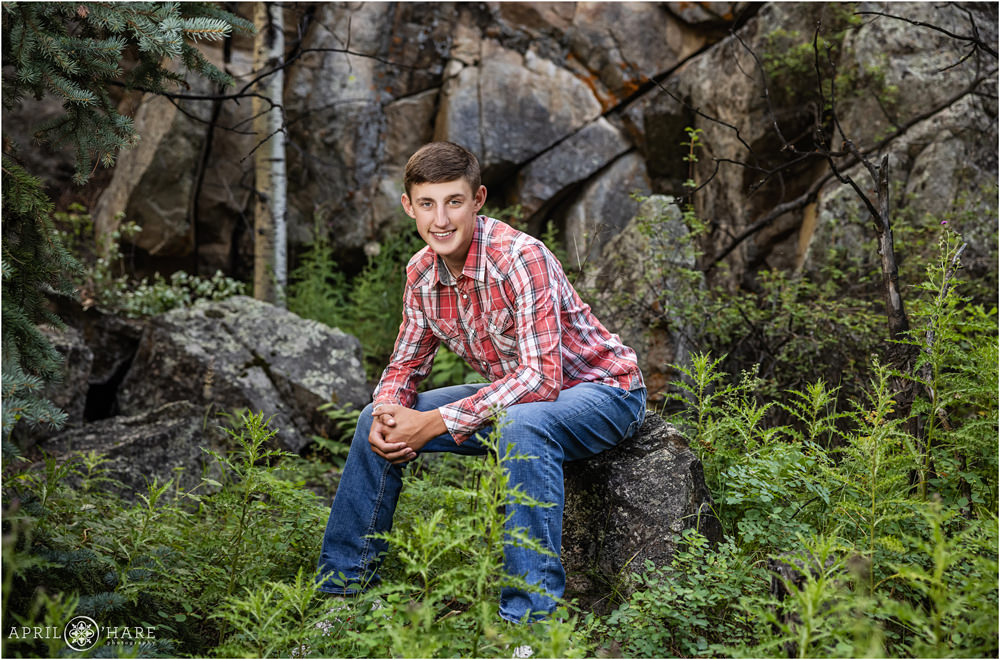 High school senior boy sitting on a large boulder in a woodsy setting in Colorado