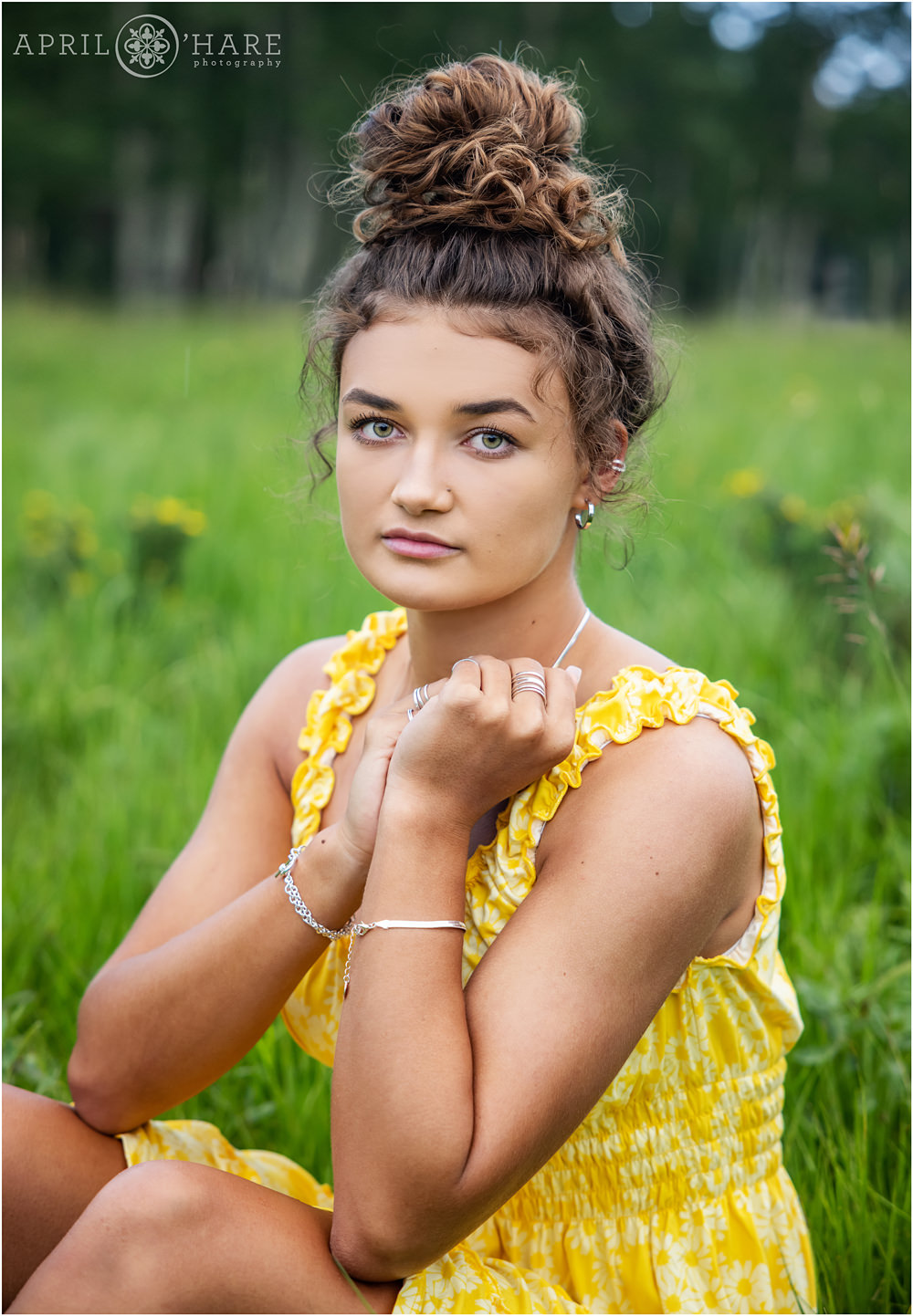 Girl with a cute messy top bun sitting in a field of green grass and yellow wildflowers wearing a pretty yellow sundress at her senior yearbook photography session in Colorado
