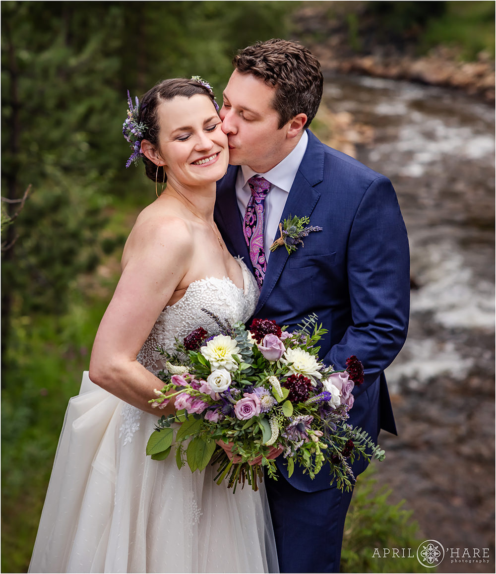 Groom kisses bride's cheek in front of Fall River on their wedding day at Estes Park Condos