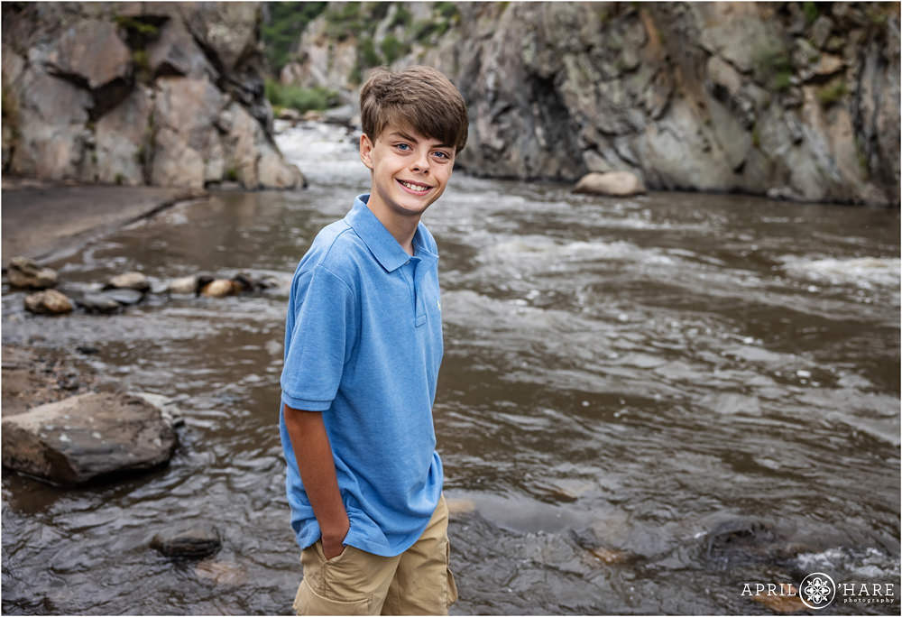 Teen boy wearing light blue shirt with khaki shorts stands in the Big Thompson River at his family's photography session at Viestenz-Smith Mountain Park