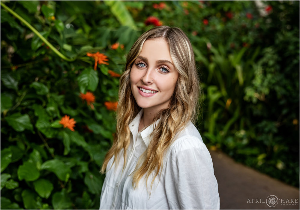 A senior girl wearing a white top stands in front of a pretty orange tropical floral plant at Denver Botanic Gardens for her Senior Photos