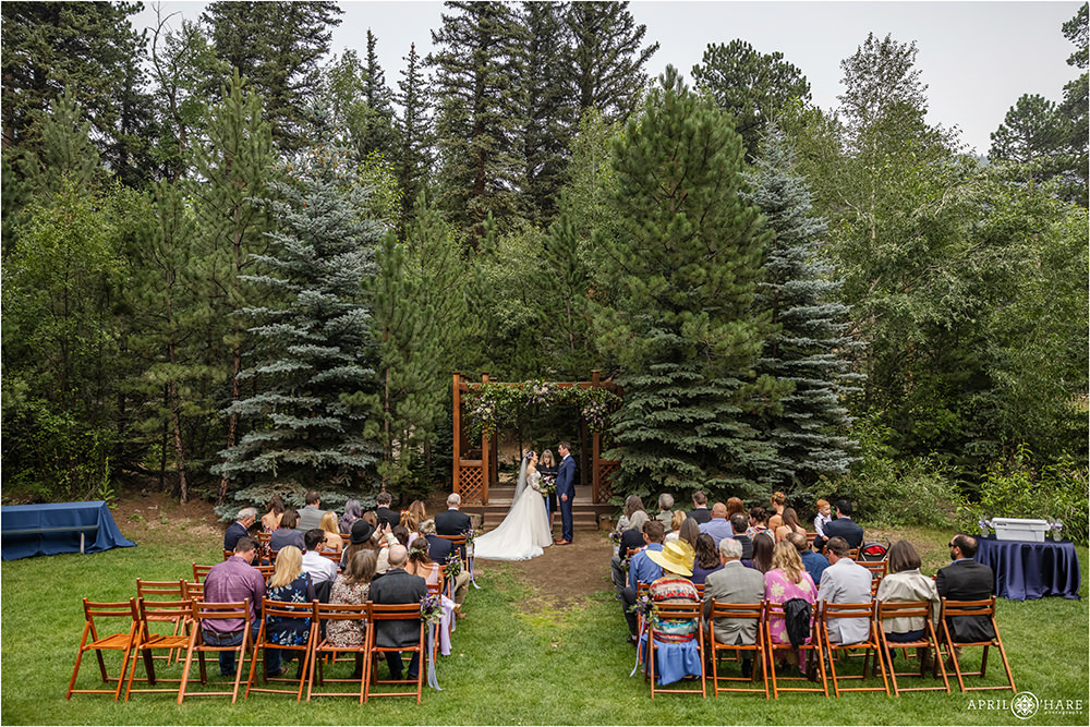 Wide angle photo of entire wedding ceremony location with bride and groom in front of a red wood gazebo decorated with purple flowers
