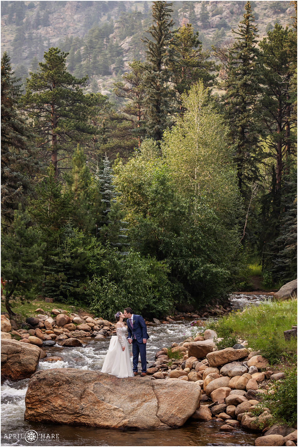 Adventurous bride and groom climb up onto a large rock in the middle of Fall River and kiss while holding hands on their wedding day at Estes Park Condos