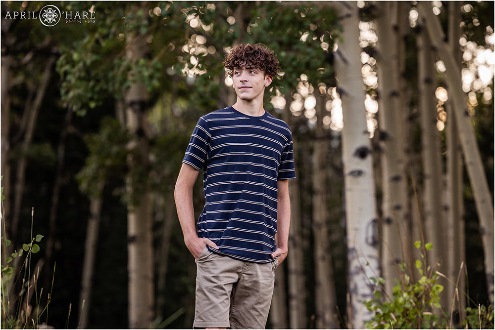 High school senior boy with curly hair and wearing a blue striped tshirt stands in a grove of Aspen Trees in Colorado