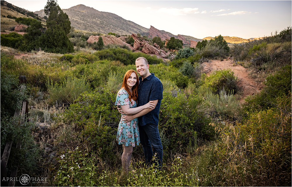 A tall man wearing a blue button down shirt poses with his red head fiance wearing a light blue floral dress during summer at East Mount Falcon Trailhead