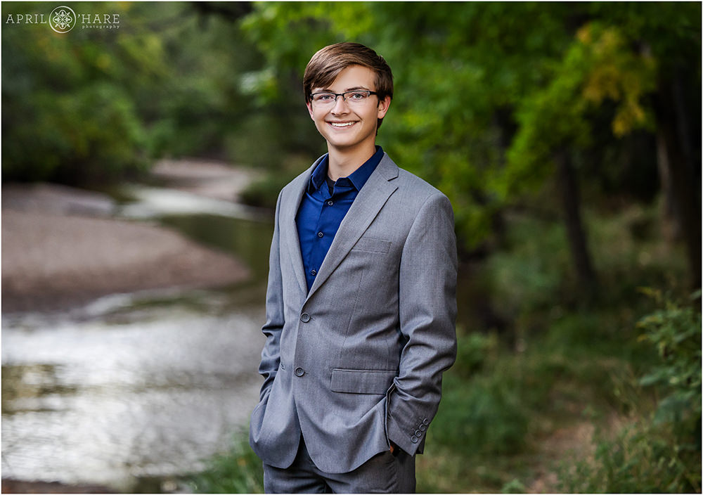 Handsome high school senior boy wearing a gray suit with a dark blue button down shirt at Cherry Creek Trail in East Denver