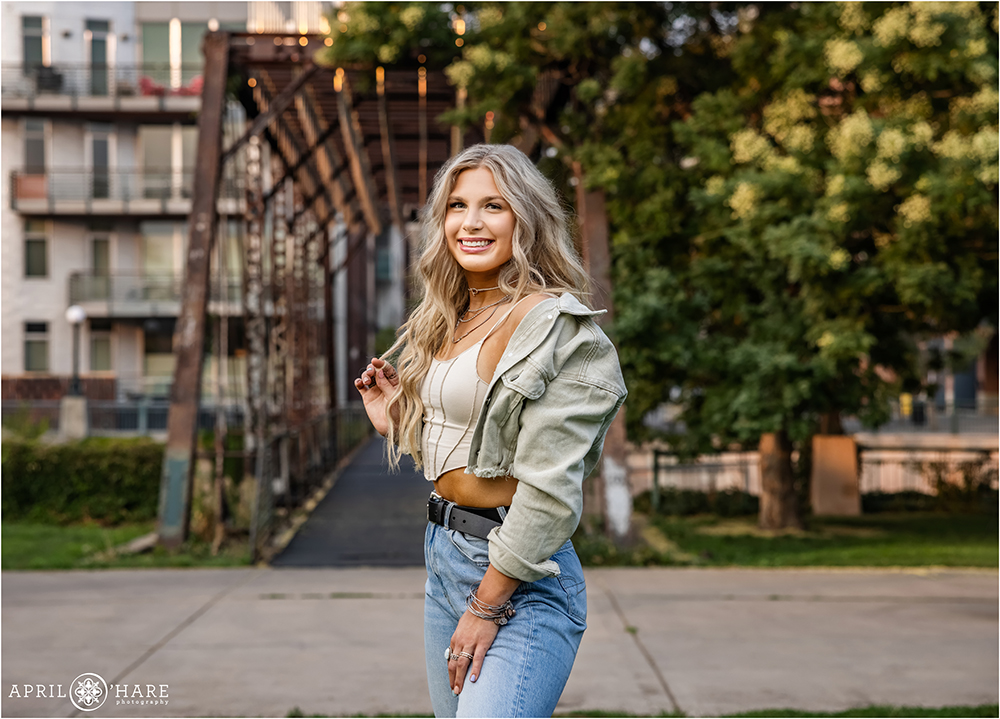 A high school senior girl with long blonde hair poses in front of a cool iron bridge along the Cherry Creek trail near downtown Denver Colorado during summer