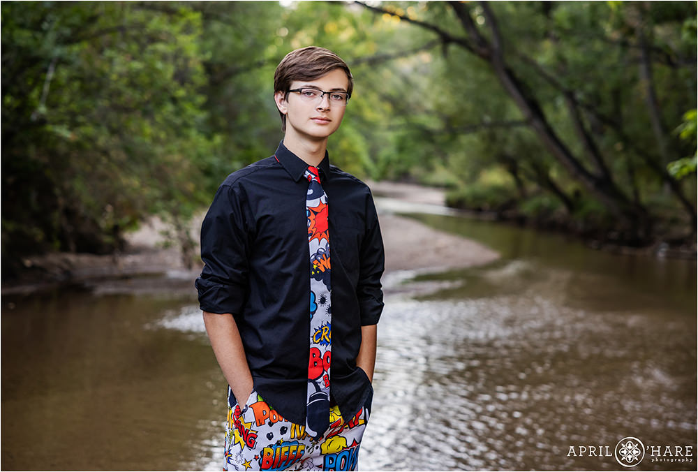 High school senior boy with serious expression on his face wearing glasses with black button down shirt with rolled up sleeves and a comic book themed tie and pants