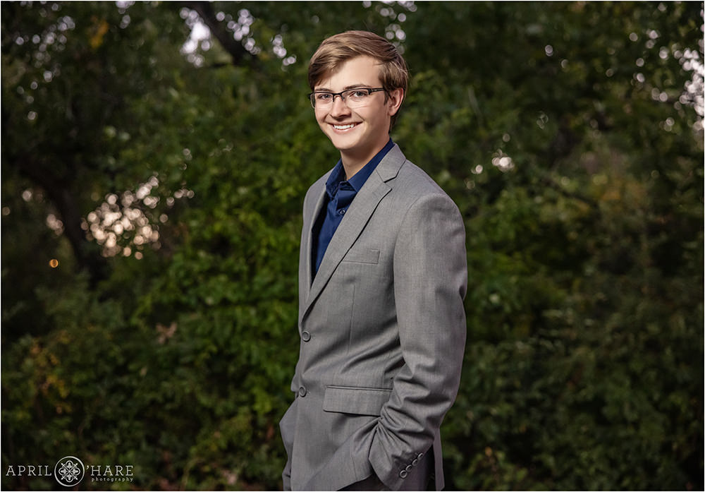 High school senior boy wearing glasses and a gray suit with a green foliage backdrop along the Cherry Creek Trail near Four Mile Historic House in East Denver