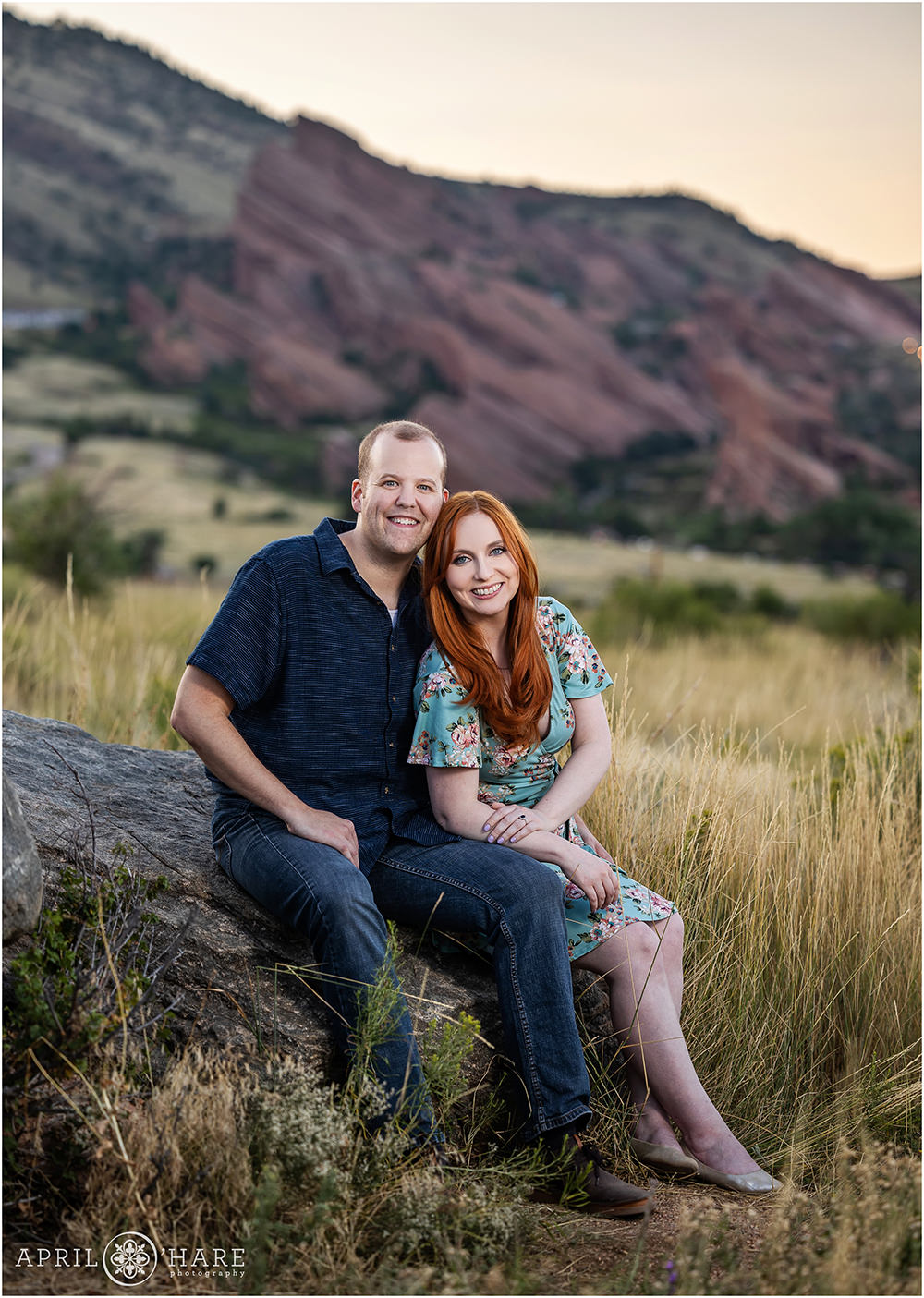 Engaged couple pose seated on a rock with Red Rocks in the backdrop at their engagement portrait session
