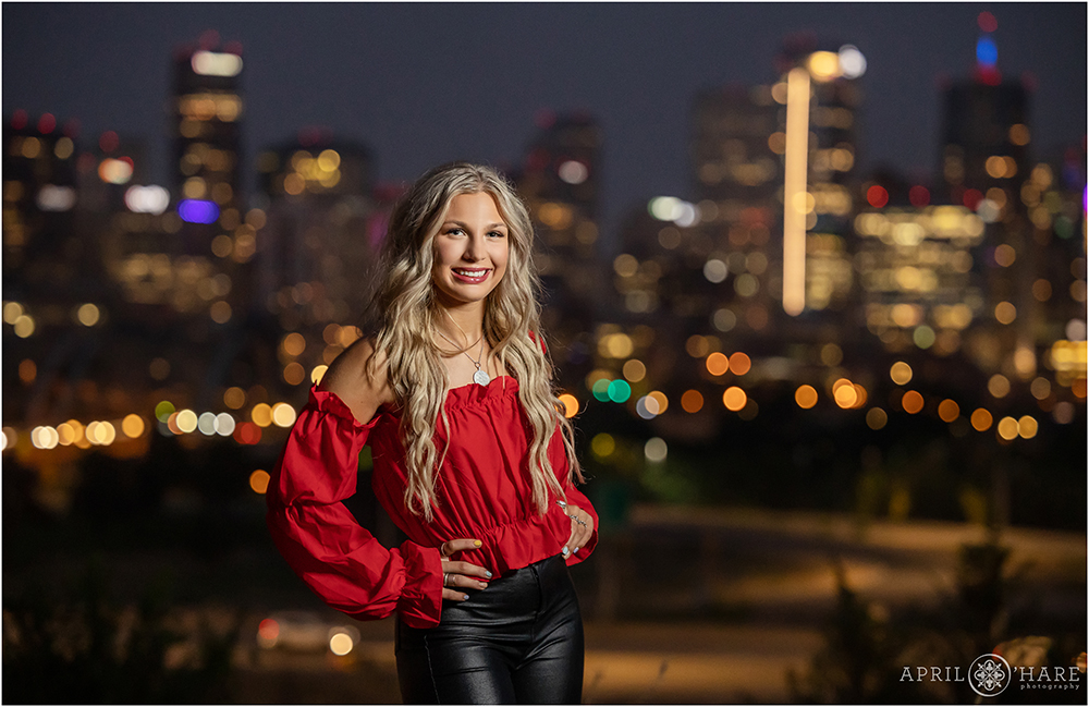 High school senior girl with long blonde hair poses in front of a glowing city skyline in Denver Colorado