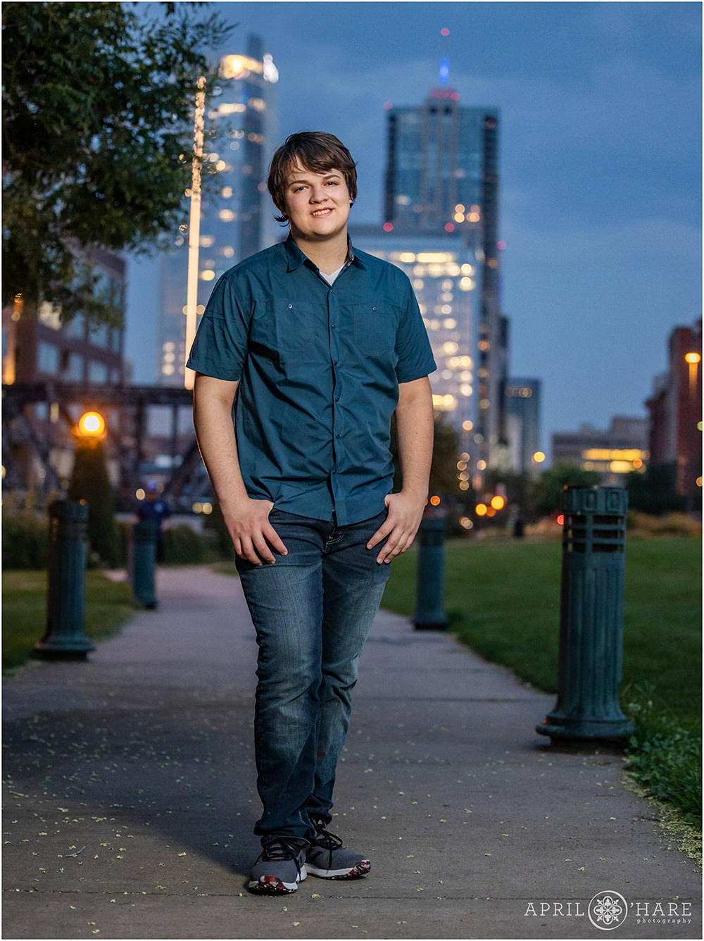 High school senior boy wearing jeans and a blue button down top with a pretty city backdrop lit up at dusk