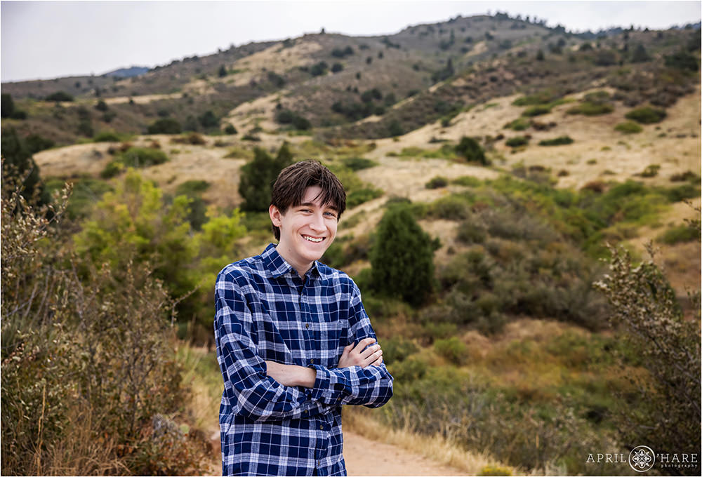 High school senior boy wearing a blue plaid button down shirt poses on a dirt trail at Mount Falcon East in Colorado