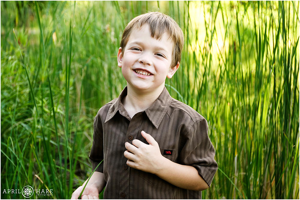 Young boy wearing a brown short sleeve button up shirt with stripes in front of tall grass