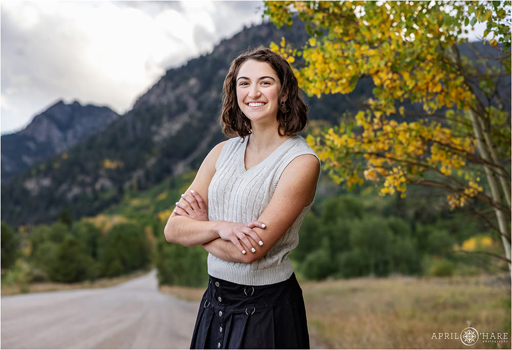 High school senior girl with shoulder length brunette hair wearing a sleeveless cream colored sweater top with a black skirt poses in front of the fall color along a dirt road in Frisco Colorado