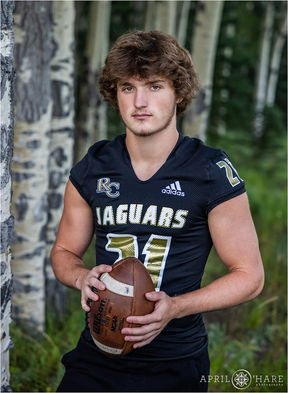 High school senior boy wearing a black football jersey and holding a wilson football stands in a grove of aspen trees for his Colorado yearbook photo